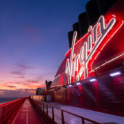 Illuminated Virgin Voyages sign on a cruise ship at sunset, with a sleek red exterior and dramatic sky in the background.