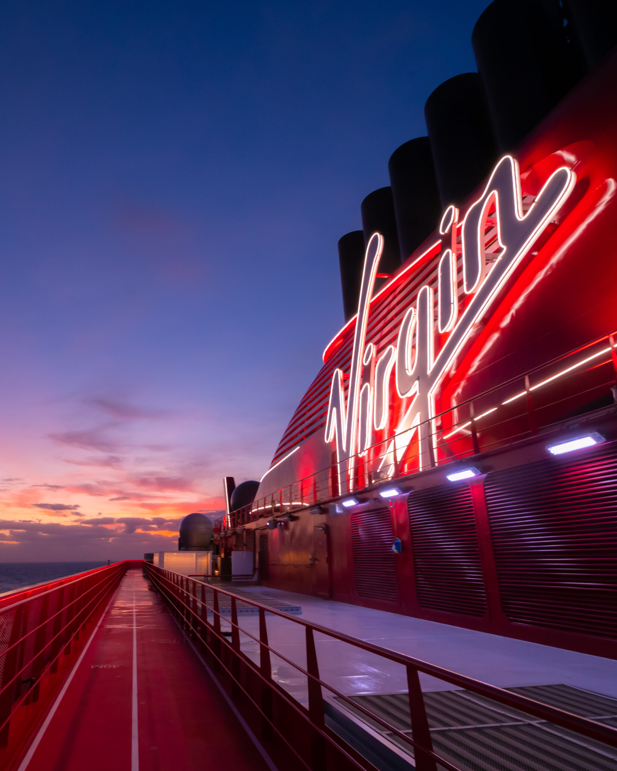 Illuminated Virgin Voyages sign on a cruise ship at sunset, with a sleek red exterior and dramatic sky in the background.