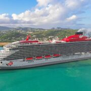 Virgin Voyages' Valiant Lady cruise ship docked in a tropical port, surrounded by turquoise waters with lush green hills and a coastal town in the background. The ship features a sleek silver and red exterior, multiple decks with glass balconies, and the signature Virgin mermaid logo on the hull. White clouds scatter across the blue sky, enhancing the scenic view.