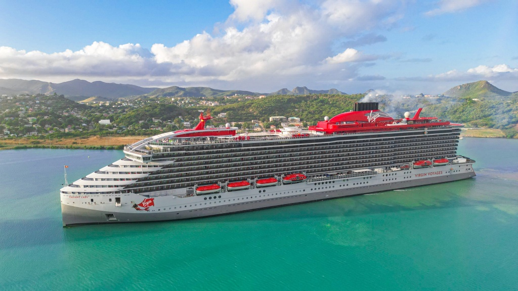 Virgin Voyages' Valiant Lady cruise ship docked in a tropical port, surrounded by turquoise waters with lush green hills and a coastal town in the background. The ship features a sleek silver and red exterior, multiple decks with glass balconies, and the signature Virgin mermaid logo on the hull. White clouds scatter across the blue sky, enhancing the scenic view.